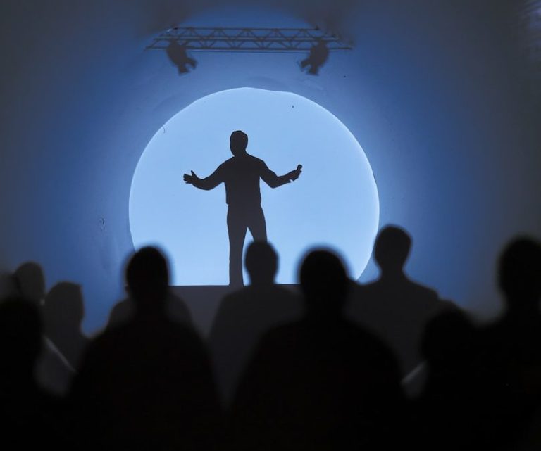 Live-Audiodeskription Eine Silhouette eines Sprechers auf einer Bühne vor einem großen, hellen Hintergrund.