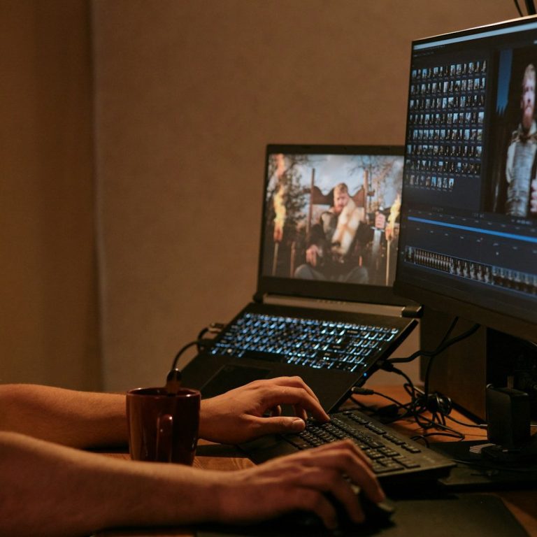 Untertitelung Person, nur Hände sichtbar, bearbeitet Video an zwei Computermonitoren, eine Tasse steht auf dem Pult.